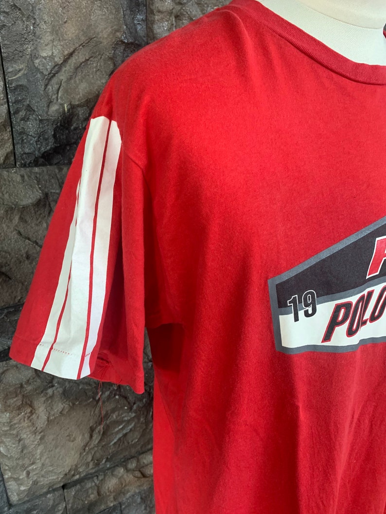 Op de afbeelding: Rood t-shirt met witte strepen op de mouw en een afbeelding op de voorkant. De afbeelding bevat het woord "POLO" en het nummer "19". Het shirt is gemaakt van een zacht materiaal.