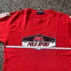 Op de afbeelding: Rood t-shirt met witte strepen op de mouwen. Het shirt heeft de tekst "RL POLO SPORT 1967" in een zwart, wit en rood ontwerp. Het shirt wordt gemeten met een meetlint.