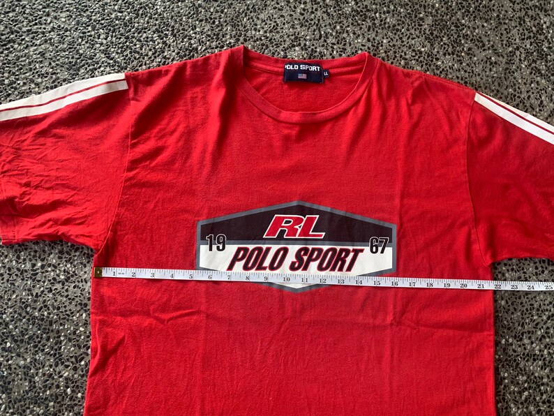 Op de afbeelding: Rood t-shirt met witte strepen op de mouwen. Het shirt heeft de tekst "RL POLO SPORT 1967" in een zwart, wit en rood ontwerp. Het shirt wordt gemeten met een meetlint.