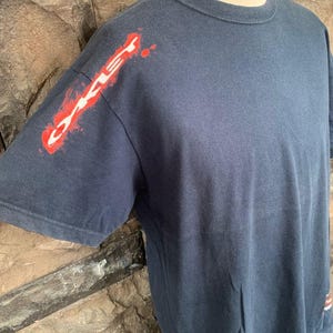 May include: A navy blue short-sleeve t-shirt with the word "Oakley" in red and white on the left sleeve. The shirt has a crew neck and a simple design, suitable for casual wear.