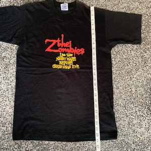 Peut inclure: T-shirt noir avec l'inscription rouge "Zthe Zombies" et "The Tour Nightmare Before Christmas Eve" en dessous. La longueur du t-shirt est d'environ 74 cm, mesur&eacute;e avec une r&egrave;gle.