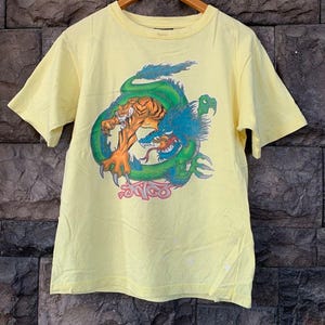 May include: Yellow t-shirt featuring a graphic of a tiger and a dragon in a battle scene. The dragon is green and blue, and the tiger is orange and black. The word "Sento" is printed in pink. The shirt is hanging on a wooden hanger.