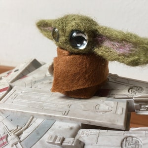 Puede incluir: Una figura de peluche hecha a mano de un personaje con ojos grandes y piel verde, que lleva una capa marrón. La figura se coloca sobre un modelo de una nave espacial, con un exterior gris detallado.