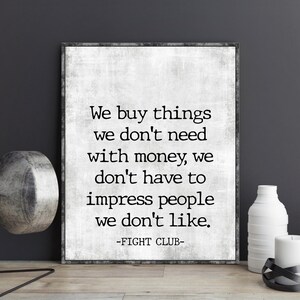 May include: Black and white typography print with a quote from the movie Fight Club: "We buy things we don't need with money, we don't have to impress people we don't like." The quote is framed in a black frame and is displayed on a wooden surface.
