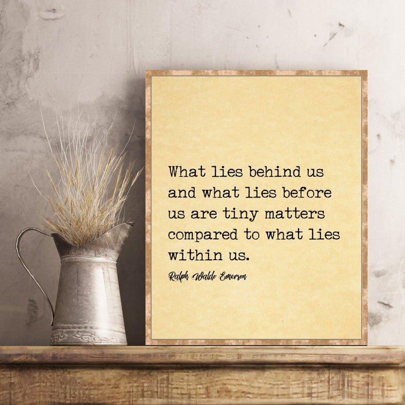 May include: A framed print with a quote by Ralph Waldo Emerson, displayed on a wooden shelf. The quote is in black text on a cream-colored background. A metal pitcher with dried grass sits to the left.
