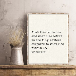 May include: A framed print with a quote by Ralph Waldo Emerson, displayed on a wooden shelf. The quote reads, "What lies behind us and what lies before us are tiny matters compared to what lies within us." A metal pitcher with dried grass sits to the left.