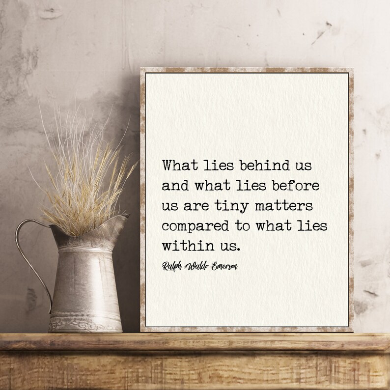 May include: A framed print with a quote by Ralph Waldo Emerson, displayed on a wooden shelf. The quote reads, "What lies behind us and what lies before us are tiny matters compared to what lies within us." A metal pitcher with dried grass sits to the left.