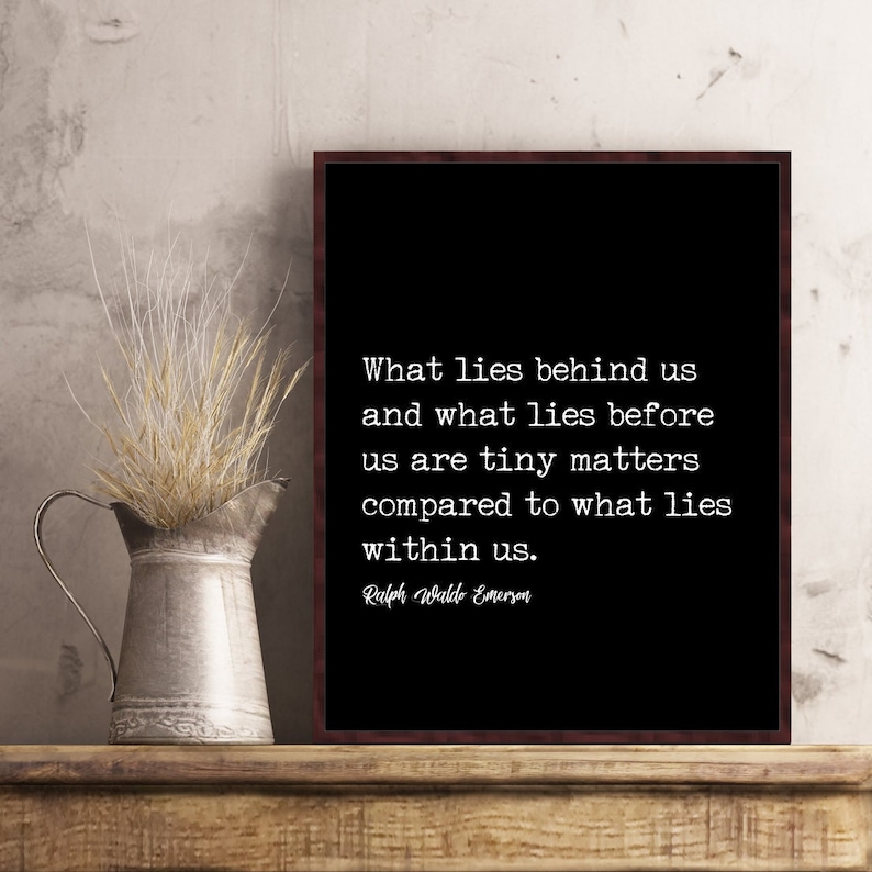 May include: A framed black print with white text quoting Ralph Waldo Emerson: "What lies behind us and what lies before us are tiny matters compared to what lies within us." A metal pitcher with dried grass sits to the left.