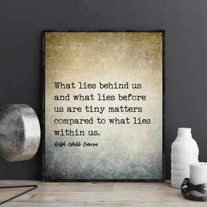 May include: Framed print with a quote by Ralph Waldo Emerson: "What lies behind us and what lies before us are tiny matters compared to what lies within us." The print has a textured, neutral background. Decorative objects, including white vases and a candle, are in the foreground.
