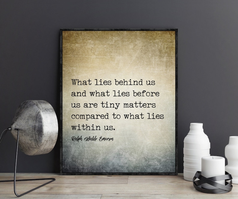 May include: Framed print with a quote by Ralph Waldo Emerson: "What lies behind us and what lies before us are tiny matters compared to what lies within us." The print has a textured, neutral background. Decorative objects, including white vases and a candle, are in the foreground.