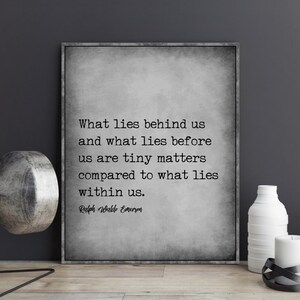 May include: A framed print with a gray background features a quote by Ralph Waldo Emerson: "What lies behind us and what lies before us are tiny matters compared to what lies within us." Decorative items are on a wooden surface.