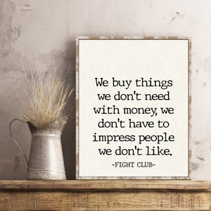 May include: A white framed print with a black and white quote from the movie Fight Club. The quote reads: "We buy things we don't need with money, we don't have to impress people we don't like." -FIGHT CLUB-