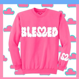 May include: A bright pink sweatshirt with the word "BLESSED" in puffy white letters across the front. A white heart is on the neckline and on the left sleeve. The background features pink clouds with blue outlines.