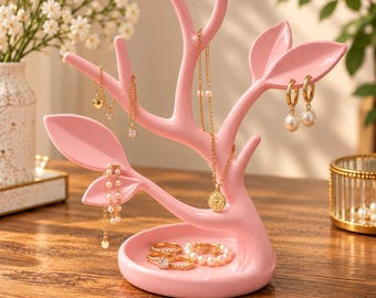 Op de afbeelding: Een lichtroze sieradenstandaard in de vorm van een boom met takken en bladeren. De basis is een ronde schaal met daarin diverse gouden ringen. Aan de takken hangen gouden kettingen, een gouden ketting met een ronde hanger en gouden oorbellen.