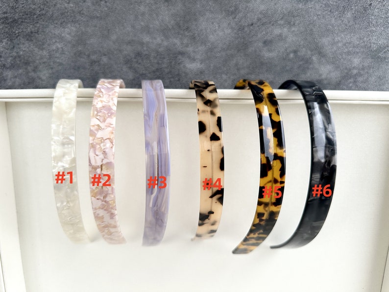 May include: Six different plastic headbands in various patterns. The headbands are numbered 1 through 6.  #1 is white with a pearlescent finish. #2 is pink with a pearlescent finish. #3 is light purple with a marbled finish. #4 is brown and white with a tortoise shell pattern. #5 is brown and black with a tortoise shell pattern. #6 is black with a marbled finish.