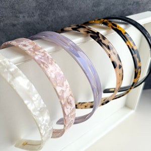 May include: Five plastic headbands in a variety of colors and patterns. The headbands are arranged in a row, with the black headband at the top and the white headband at the bottom. The headbands are all about 1 inch wide.