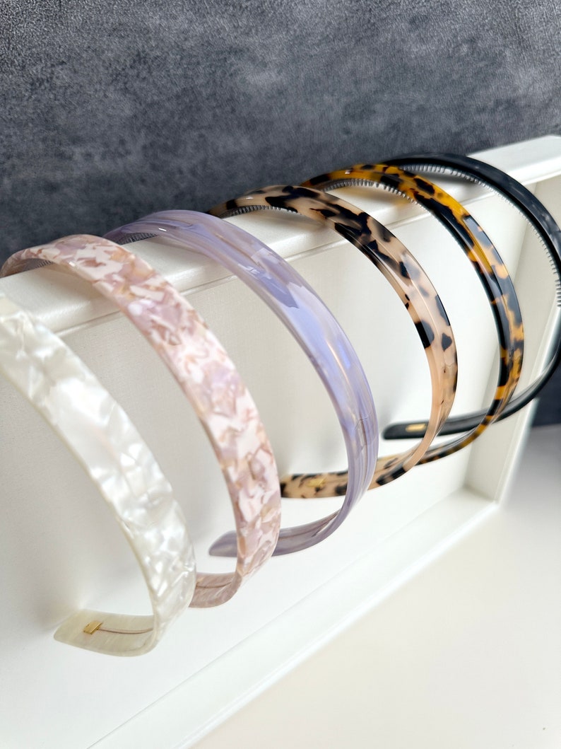 May include: Five plastic headbands in a variety of colors and patterns. The headbands are arranged in a row, with the black headband at the top and the white headband at the bottom. The headbands are all about 1 inch wide.
