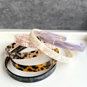 May include: A set of five hair headbands in different colors and patterns. The headbands are made of a hard plastic material and have a smooth, glossy finish. The colors include black, brown, white, and purple.