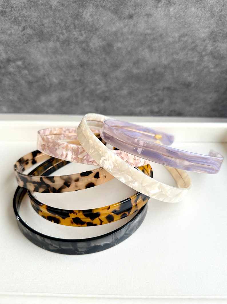 May include: A set of five hair headbands in different colors and patterns. The headbands are made of a hard plastic material and have a smooth, glossy finish. The colors include black, brown, white, and purple.