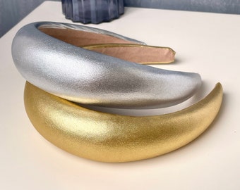 May include: Two wide headbands, one silver and one gold, made of a soft, padded material. The headbands are lying on a white surface.