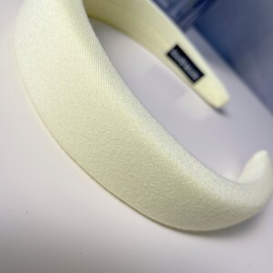 May include: A wide, off-white headband made of a soft, textured fabric. The headband is curved and has a smooth, polished finish.