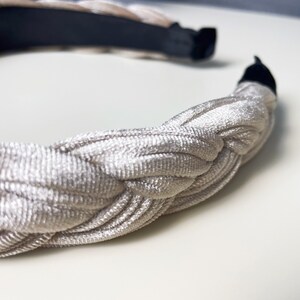 May include: A braided headband made of a soft, light beige fabric. The headband is designed to be worn around the head and has a simple, elegant look.