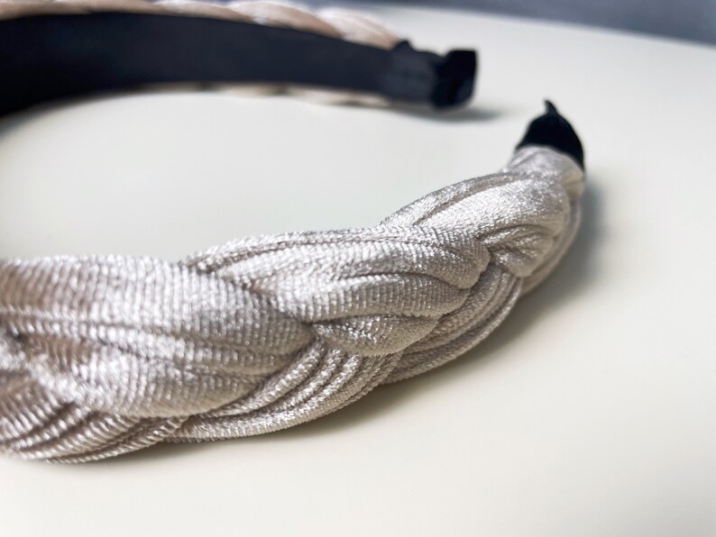 May include: A braided headband made of a soft, light beige fabric. The headband is designed to be worn around the head and has a simple, elegant look.