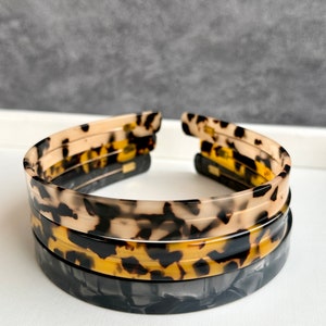May include: A set of three tortoise shell hair headbands. The headbands are stacked on top of each other. The top headband is a light brown with black and white spots. The middle headband is a dark brown with black and yellow spots. The bottom headband is black with white and gray swirls.