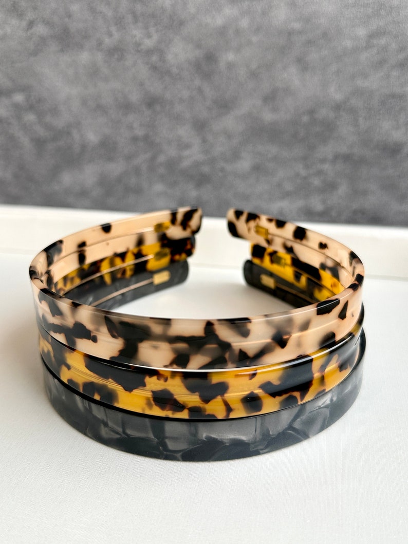 May include: A set of three tortoise shell hair headbands. The headbands are stacked on top of each other. The top headband is a light brown with black and white spots. The middle headband is a dark brown with black and yellow spots. The bottom headband is black with white and gray swirls.
