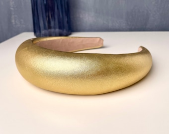 May include: A gold-colored headband made of a soft, padded material. The headband is curved and has a smooth, satin-like finish.
