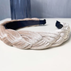 May include: A braided headband made of a soft, light beige velvet material. The headband is designed to be worn around the head and has a simple, elegant style.
