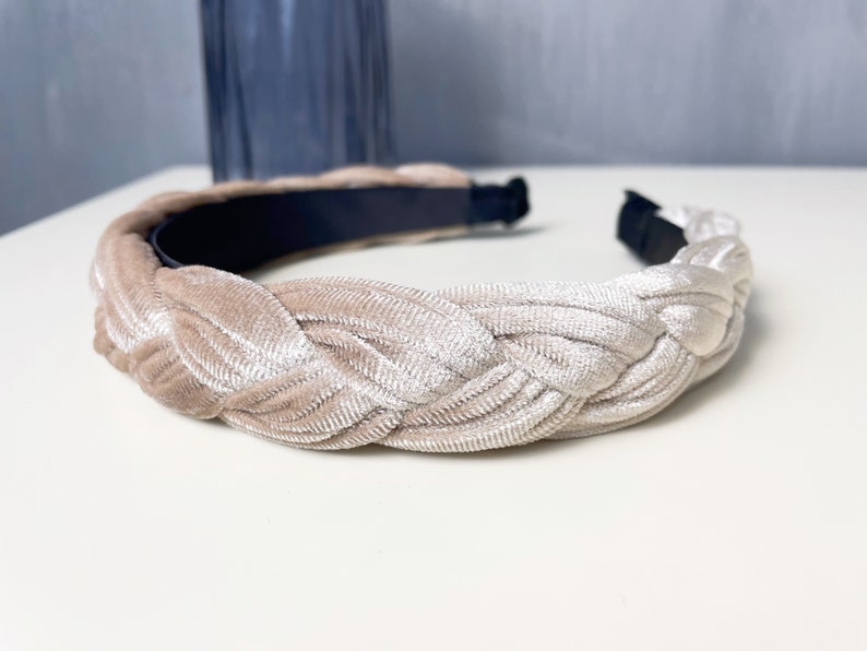 May include: A braided headband made of a soft, light beige velvet material. The headband is designed to be worn around the head and has a simple, elegant style.