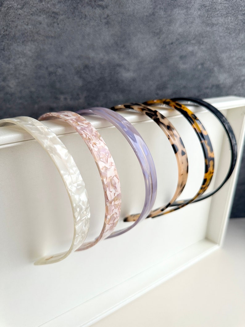 May include: A set of six plastic headbands in various colors and patterns, including white, pink, purple, brown, and leopard print. The headbands are displayed on a white surface.