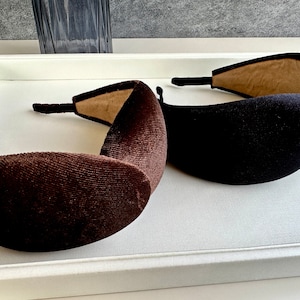 May include: Two velvet headbands, one brown and one black, with a soft, padded interior. The headbands are displayed on a white surface.