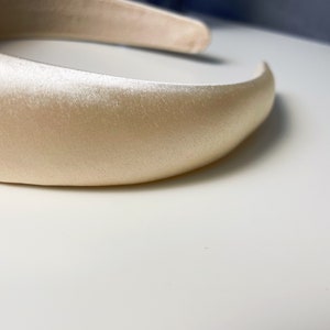 K&ouml;nnte beinhalten: Ein breites, cremefarbenes Satin-Haarband. Das Haarband ist glatt und hat einen leichten Glanz.