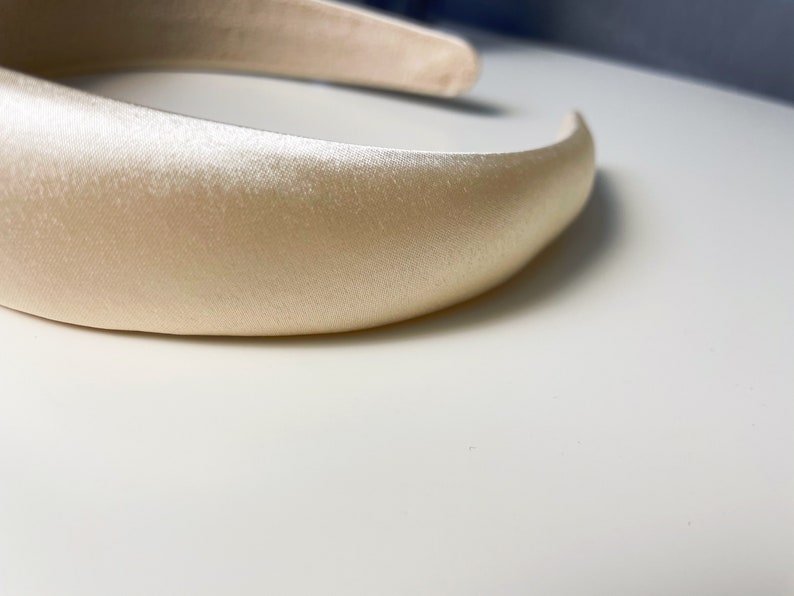 K&ouml;nnte beinhalten: Ein breites, cremefarbenes Satin-Haarband. Das Haarband ist glatt und hat einen leichten Glanz.