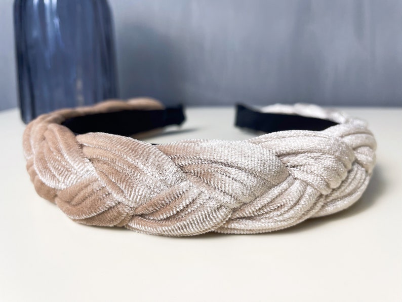 May include: A braided headband made of a soft, light beige velvet fabric. The headband is designed to be worn around the head and has a simple, elegant style.