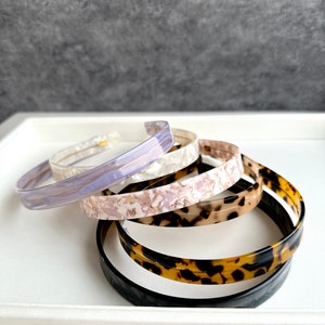 May include: Four plastic hair headbands in different patterns. The headbands are stacked on top of each other. The top headband is a light purple with a marbled pattern. The second headband is a white with a marbled pattern. The third headband is a light brown with a marbled pattern. The bottom headband is a dark brown with a tortoise shell pattern.
