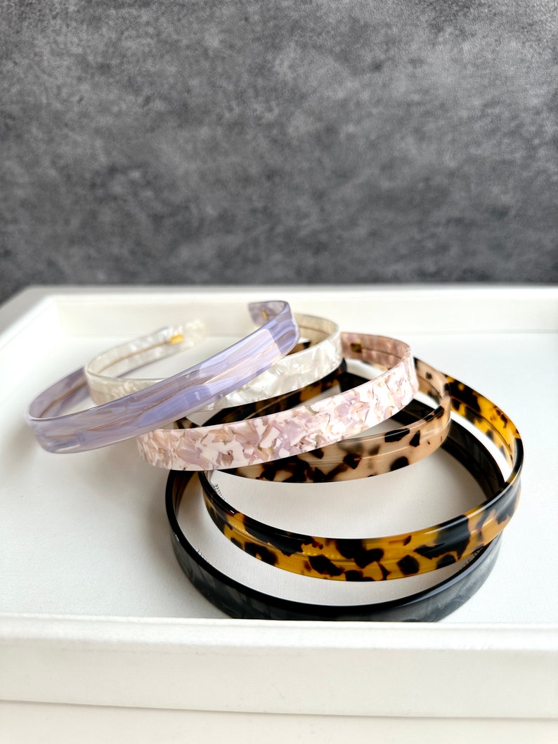 May include: Four plastic hair headbands in different patterns. The headbands are stacked on top of each other. The top headband is a light purple with a marbled pattern. The second headband is a white with a marbled pattern. The third headband is a light brown with a marbled pattern. The bottom headband is a dark brown with a tortoise shell pattern.