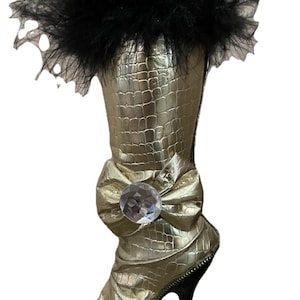 May include: A gold-colored boot with a textured crocodile-skin pattern, featuring a black feathered trim at the top opening. The boot is adorned with a large gold bow at the ankle, centered with a clear, faceted circular crystal embellishment.