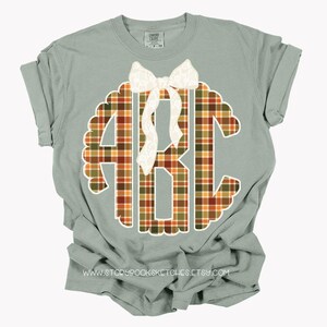 Puede incluir: Camiseta verde salvia con un monograma ABC a cuadros en naranja, marr&oacute;n y verde. Un lazo blanco adorna la parte superior del monograma. La camiseta tiene las mangas dobladas. Se ve el sitio web www.storybooksketches.etsy.com.