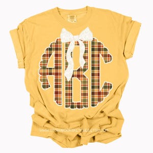 Puede incluir: Camiseta de color amarillo mostaza con un dise&ntilde;o grande con bordes festoneados que presenta las letras "ABC" en un patr&oacute;n de cuadros rojos, verdes y marrones. Un lazo blanco est&aacute; centrado encima de las letras. La direcci&oacute;n del sitio web es visible en la parte inferior.