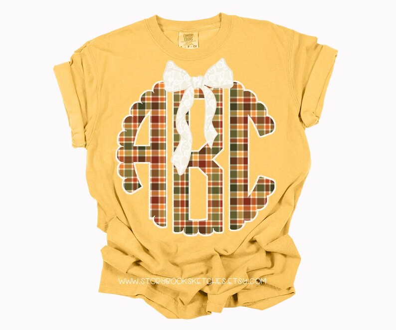 Puede incluir: Camiseta de color amarillo mostaza con un dise&ntilde;o grande con bordes festoneados que presenta las letras "ABC" en un patr&oacute;n de cuadros rojos, verdes y marrones. Un lazo blanco est&aacute; centrado encima de las letras. La direcci&oacute;n del sitio web es visible en la parte inferior.