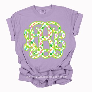 May include: A lilac-colored t-shirt featuring a large monogram design in green with a white fill. The monogram is filled with colorful shamrocks, rainbows, and hearts. The text "WWW.STORYBROOKSKETCHES.ETSY.COM" is printed at the bottom.