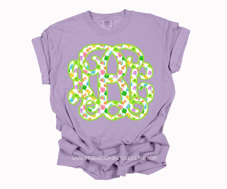 May include: A lilac-colored t-shirt featuring a large monogram design in green with a white fill. The monogram is filled with colorful shamrocks, rainbows, and hearts. The text "WWW.STORYBROOKSKETCHES.ETSY.COM" is printed at the bottom.