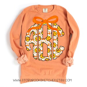 K&ouml;nnte beinhalten: Ein pfirsichfarbener Pullover mit einem Softball-Monogramm. Das Monogramm hat die Form eines Kreises mit einer Schleife oben. Die Buchstaben sind wei&szlig; mit einem Softball-Muster.
