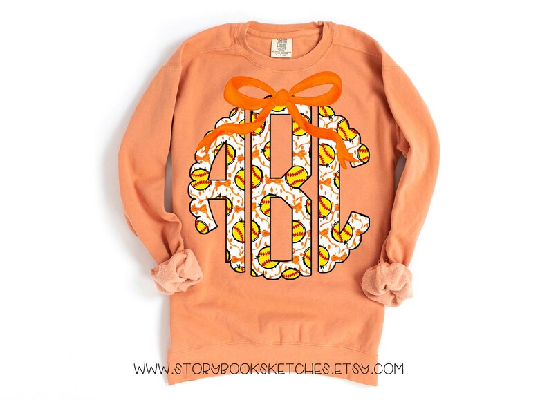 K&ouml;nnte beinhalten: Ein pfirsichfarbener Pullover mit einem Softball-Monogramm. Das Monogramm hat die Form eines Kreises mit einer Schleife oben. Die Buchstaben sind wei&szlig; mit einem Softball-Muster.