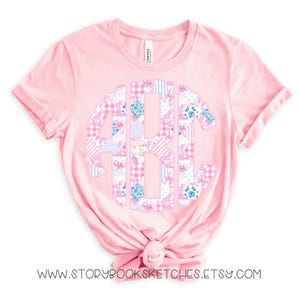 May include: A pink t-shirt with a large, white, blue, and pink monogram in the center. The monogram is made up of patchwork fabric with floral and gingham patterns. The shirt is made of a soft, comfortable material and is perfect for everyday wear.