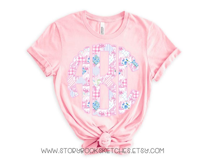 May include: A pink t-shirt with a large, white, blue, and pink monogram in the center. The monogram is made up of patchwork fabric with floral and gingham patterns. The shirt is made of a soft, comfortable material and is perfect for everyday wear.