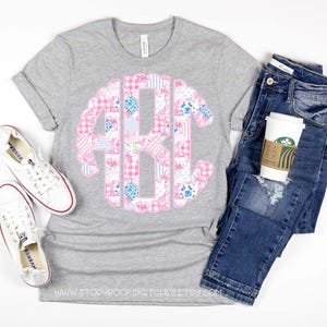 May include: A gray t-shirt with a pink and blue patchwork monogram design. The monogram is in the center of the shirt. The shirt is laying on a white surface with a pair of white sneakers and a pair of blue jeans.